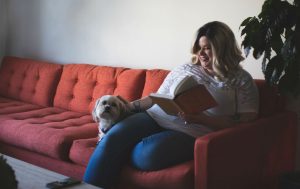 Woman sitting on red couch reaching book and petting small dog beside her