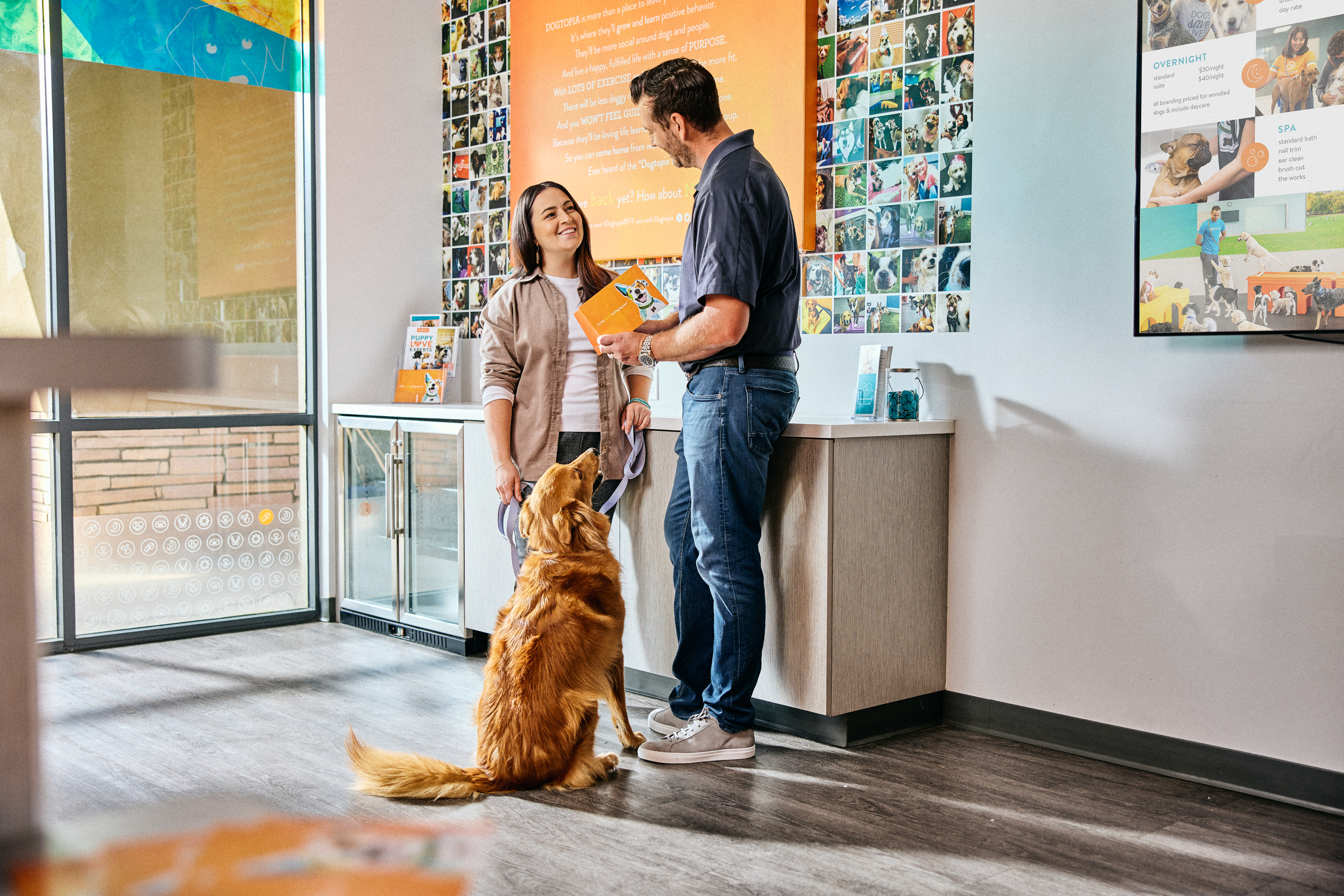 Man and woman inside Dogtopia daycare lobby with golden lab sitting and looking up at them.