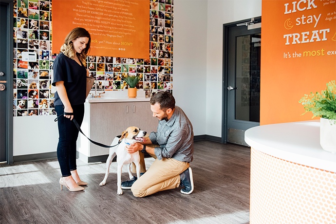 A man kneeling down to pet a dog that is on a leash, which is held by a woman in a Dogtopia lobby.
